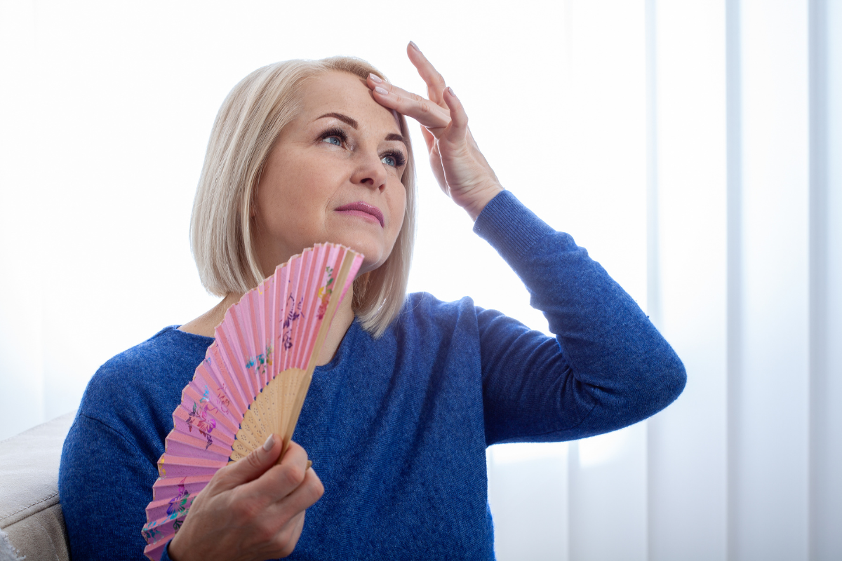 A woman sits indoors, she holds a pink floral hand fan in one hand and raises her other hand to her forehead to cool herself. She represents one of the 12 common symptoms of perimenopause, hot flashes and night sweats.