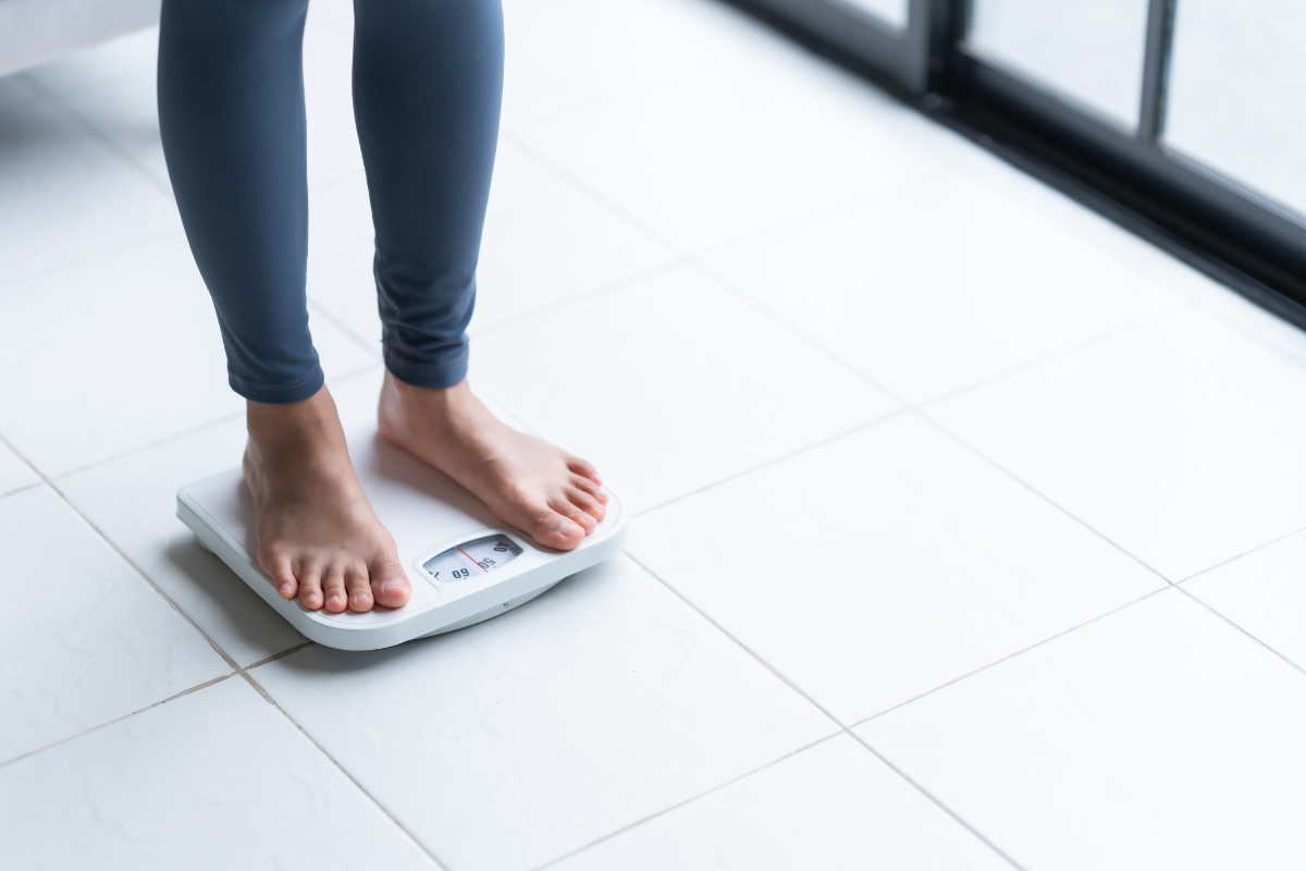image shows womans legs from the knee doown wearing grey leggings and bare feet standing on white weighing scales. the floor surface is tiled and white.
